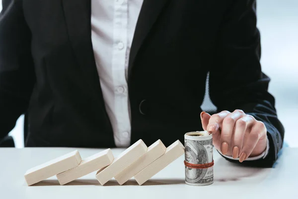 Partial view of businesswoman sitting at table with fallen row of wooden blocks and money roll — Stock Photo