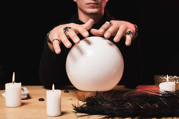 Cropped view of man holding hands above crystal ball near candles isolated on black — Stock Photo