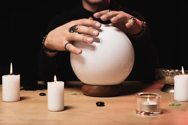 Vista recortada de las manos esotéricas cogidas por encima de la bola de cristal cerca de velas aisladas en negro - foto de stock