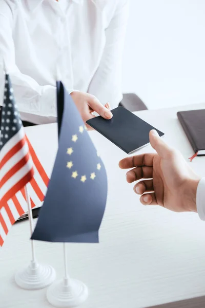 Vista cortada de agente de viagens feminino dando passaporte para turista perto de bandeiras americanas e europeias isoladas em branco — Fotografia de Stock