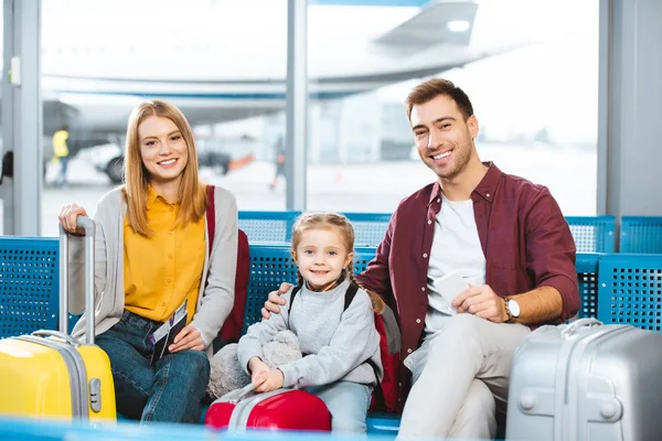 Familia feliz sentado en la sala de salida y sonriendo cerca de equipaje en el aeropuerto - foto de stock