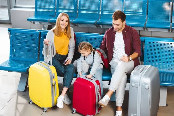 Familia feliz sentado cerca de equipaje y sonriendo mientras espera el vuelo en el aeropuerto - foto de stock