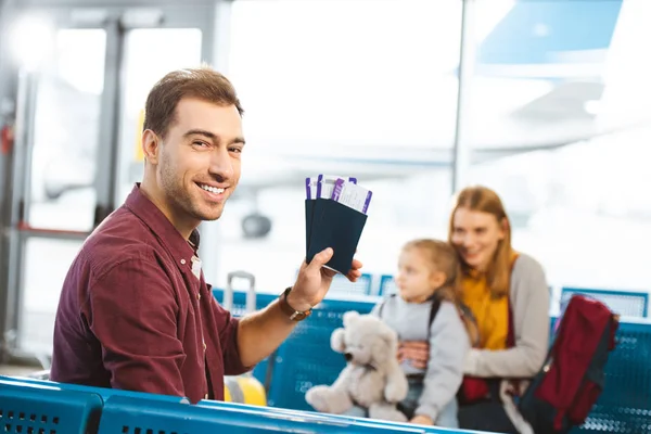 Selective focus of handsome man smiling while holding passports with air tickets with wife and daughter on background — Stock Photo