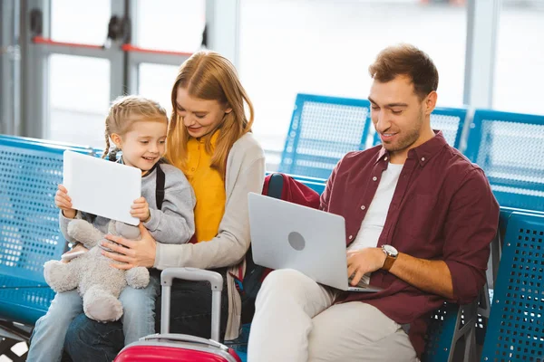 Lindo niño sosteniendo la tableta digital cerca de la madre y mirando a padre usando el ordenador portátil en aeropuerto - foto de stock