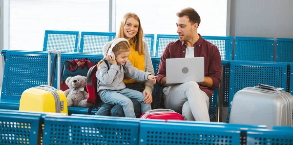 Nette Tochter zeigt mit dem Finger auf Laptop, während sie in der Nähe von Vater und Mutter auf dem Flughafen sitzt — Stockfoto