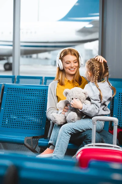 Attractive mother in headphones looking at daughter with teddy bear in departure lounge — Stock Photo