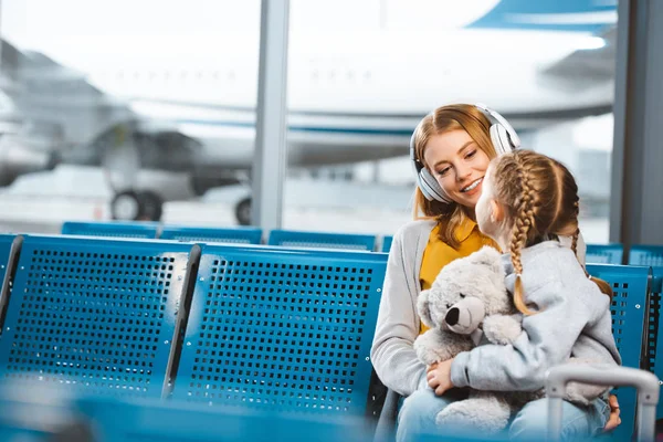 Schöne Mutter mit Kopfhörern schaut Tochter mit Teddybär in Abflughalle an — Stockfoto
