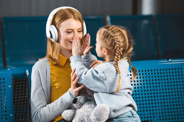 Hija tocando cara de hermosa madre en auriculares en el aeropuerto - foto de stock
