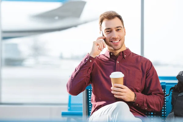 Cheerful man talking on smartphone and holding paper cup in hand in airport — Stock Photo