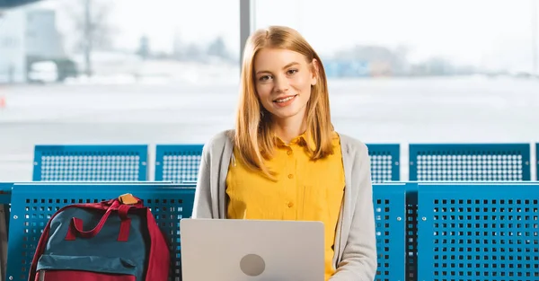 Attraktive Frau lächelt, während sie Laptop in der Abflughalle benutzt — Stockfoto