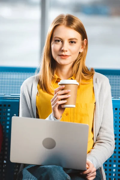 Atractiva mujer sentada con el ordenador portátil y sosteniendo la taza de papel en la sala de salida del aeropuerto - foto de stock