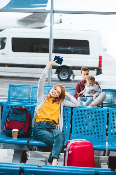 Selektiver Fokus einer lächelnden Frau mit Pass über dem Kopf im Flughafen in der Nähe eines Rucksacks mit Menschen im Hintergrund — Stockfoto