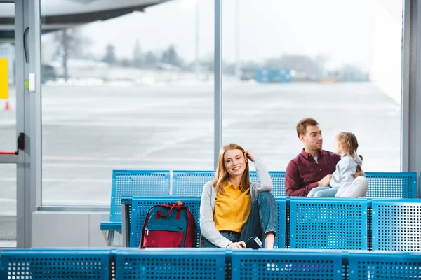 Enfoque selectivo de la mujer sonriente sentada en el aeropuerto cerca de la mochila con la gente en el fondo - foto de stock