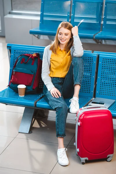 Atractiva mujer sentada en la sala de espera del aeropuerto cerca de la mochila y el equipaje - foto de stock