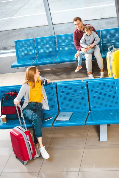 Feliz padre sentado con su hija en el aeropuerto cerca de la mujer con el equipaje - foto de stock