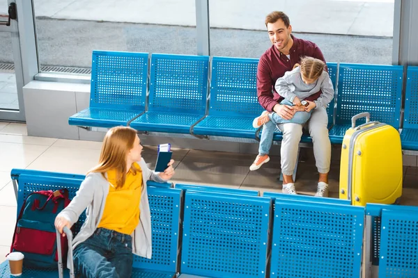 Padre guapo sentado con hija en el aeropuerto cerca de la mujer con el equipaje - foto de stock