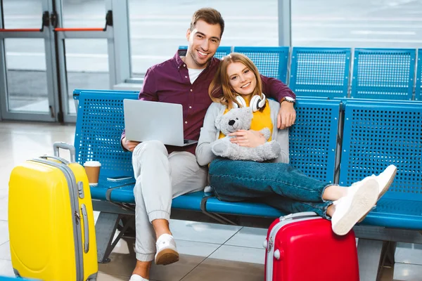 Novio feliz con portátil sonriendo con la novia sosteniendo oso de peluche en el aeropuerto - foto de stock