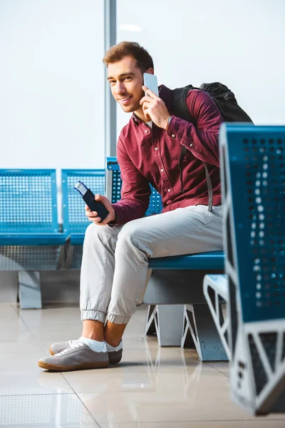 Hombre alegre hablando en el teléfono inteligente y sonriendo mientras está sentado en la sala de espera - foto de stock
