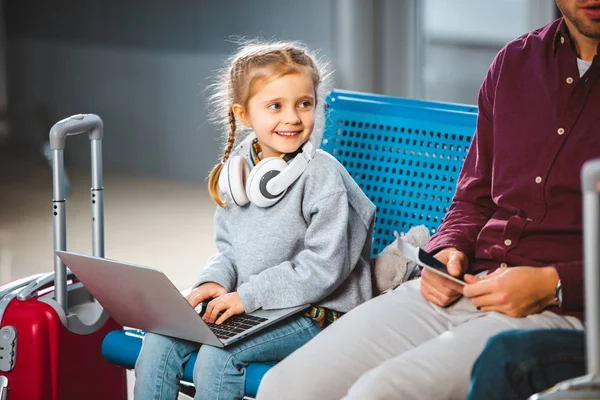 Niño sonriente en los auriculares usando el ordenador portátil cerca de papá en el aeropuerto - foto de stock
