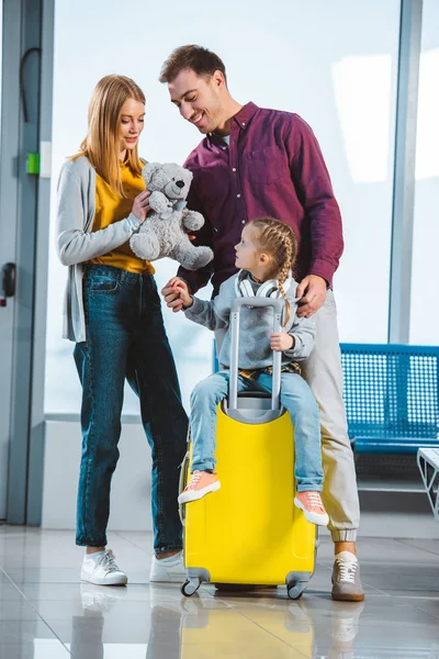 Adorable niño en auriculares sentados en la maleta mientras madre sosteniendo oso de peluche y de pie cerca del marido en la sala de salida - foto de stock