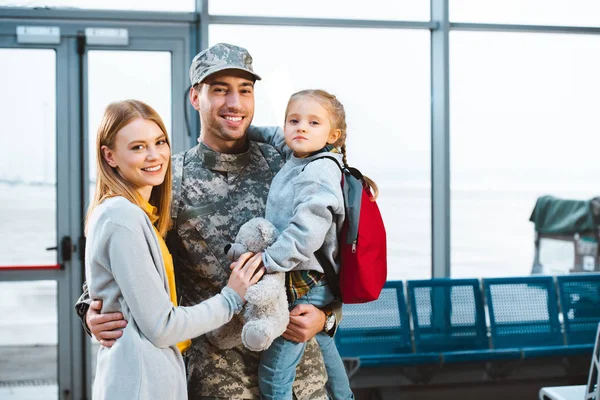 Весёлый отец в военной форме держит в руках милую дочь и стоит рядом с женой в аэропорту — стоковое фото