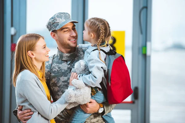 Sonriente padre en uniforme militar sosteniendo en brazos hija cerca de esposa en aeropuerto - foto de stock