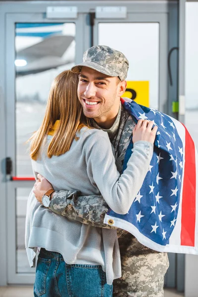 Vista posteriore della donna che abbraccia sorridente fidanzato in uniforme militare con bandiera americana in aeroporto — Foto stock