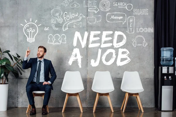 Businessman in suit showing idea gesture and sitting in waiting hall with need a job lettering on wall — Stock Photo