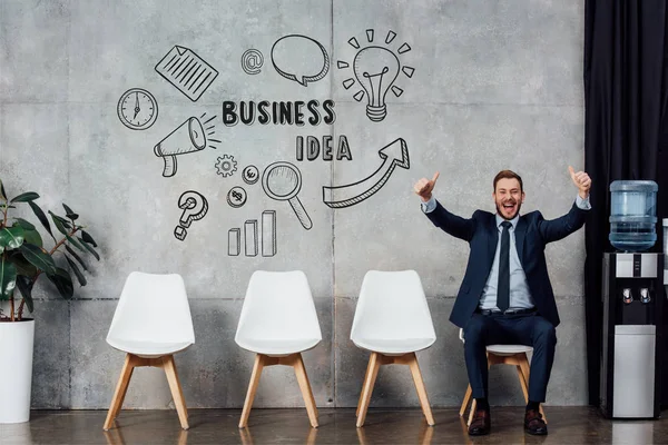 Excited businessman in suit sitting and showing thumbs up in waiting hall with business idea lettering on wall — Stock Photo