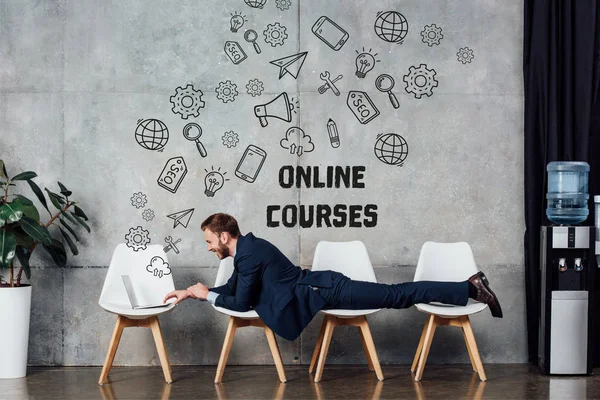Businessman lying on chairs and using laptop in waiting hall with online courses lettering on wall — Stock Photo