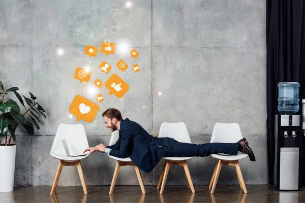 Businessman lying on chairs and using laptop in waiting hall with social media icons on wall — Stock Photo