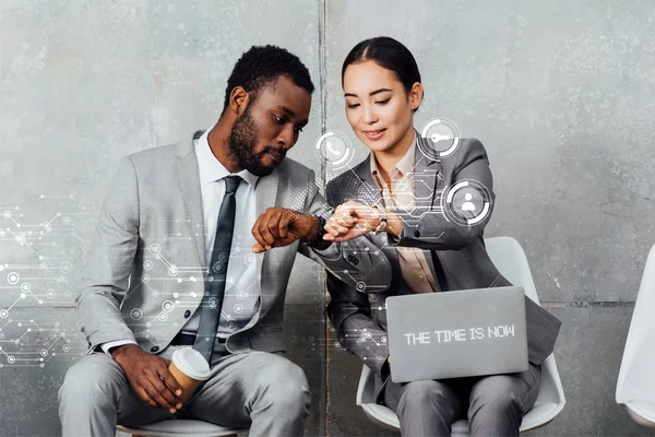 Multiethnic colleagues with laptop and coffee sitting and looking at smartwatches in waiting hall with the time is now lettering on laptop and notifications icons — Stock Photo