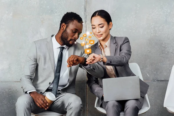 Multiethnic colleagues with laptop and coffee sitting and looking at smartwatches in waiting hall with social media notifications icons — Stock Photo