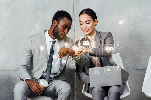 Multiethnic colleagues with laptop and coffee sitting and looking at smartwatches in waiting hall with notifications icons — Stock Photo