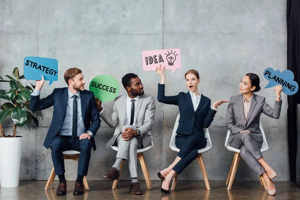 Empresarios multiétnicos felices sosteniendo burbujas del discurso con la idea, el éxito, la planificación y las letras de la estrategia mientras que se sienta en la sala de espera - foto de stock