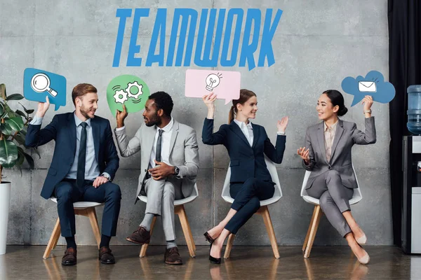Empresarios multiétnicos felices sosteniendo burbujas de habla con iconos de trabajo en equipo mientras están sentados en la sala de espera - foto de stock