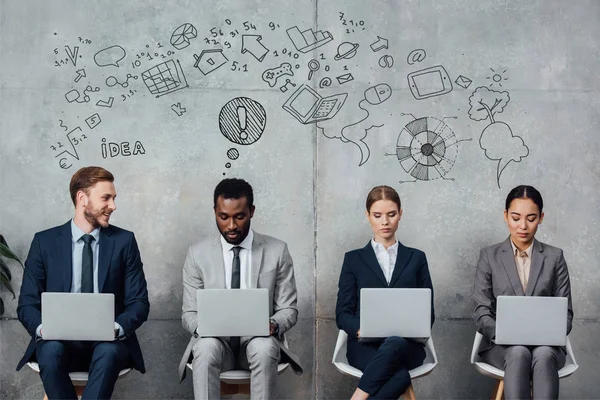 Empresarios multiétnicos sentados en sillas y usando computadoras portátiles en la sala de espera con iconos de ideas en la pared - foto de stock