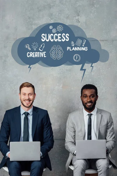 Hombres de negocios multiétnicos sonrientes mirando a la cámara y usando computadoras portátiles en la sala de espera con ilustración de burbujas de habla con letras creativas, exitosas y de planificación - foto de stock