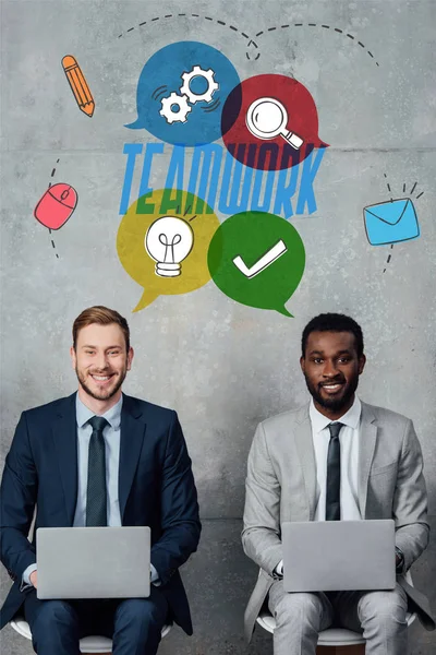 Hombres de negocios multiétnicos sonrientes mirando a la cámara y usando computadoras portátiles en la sala de espera con letras de trabajo en equipo en la pared - foto de stock