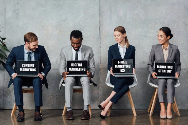Smiling multiethnic businesspeople sitting on chairs and holding laptops with content, digital, e-mail marketing and marketing strategy lettering on screens in waiting hall — Stock Photo