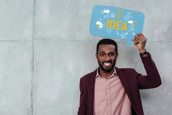 Sonriente afroamericano casual hombre de negocios mirando a la cámara y sosteniendo la burbuja del habla con letras de la idea - foto de stock