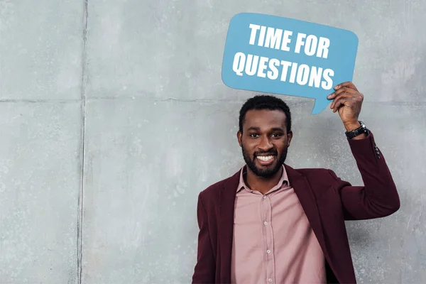 Sonriente afroamericano casual hombre de negocios mirando a la cámara y sosteniendo la burbuja del habla con el tiempo para las preguntas letras - foto de stock