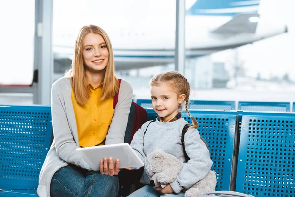 Schöne Mutter hält digitales Tablet in der Hand und sitzt mit Tochter im Flughafen — Stockfoto