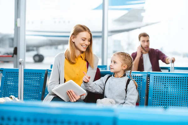Селективное внимание матери, держащей цифровой планшет и улыбающейся, глядя на дочь в аэропорту — стоковое фото