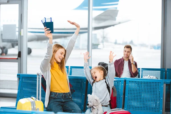 Selektiver Fokus glückliche Mutter und Tochter halten die Hände über dem Kopf und lächeln im Flughafen mit Mann im Hintergrund — Stockfoto