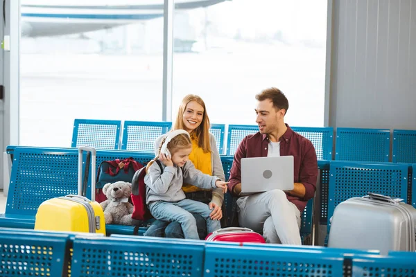 Adorable hija apuntando con el dedo a la computadora portátil mientras está sentado cerca de papá y mamá en el aeropuerto - foto de stock