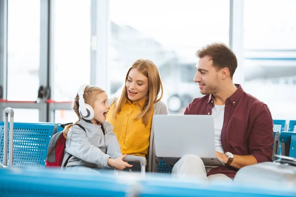 Aufgeregtes Kind mit Kopfhörern schaut Vater an, während es neben Mutter im Flughafen sitzt — Stockfoto