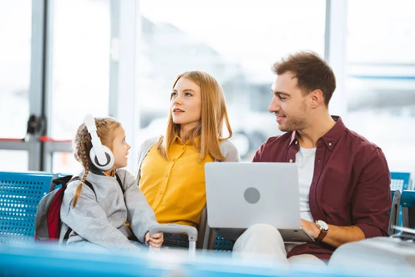 Cute kid in headphones looking at mother while sitting near father in airport — Stock Photo