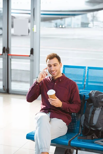 Hombre feliz hablando en el teléfono inteligente y sosteniendo la taza de papel en la mano en el aeropuerto - foto de stock