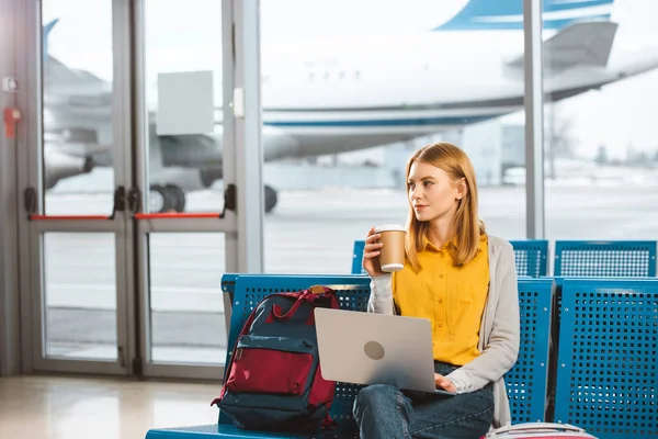 Hermosa mujer sentada con el ordenador portátil y la celebración de taza desechable en la mano en el aeropuerto - foto de stock